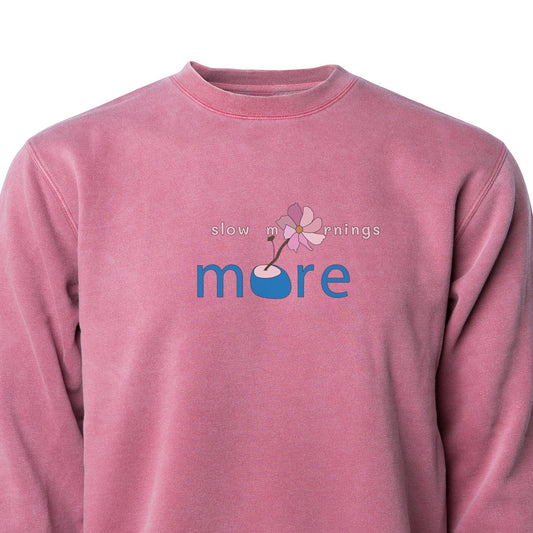A pink crew neck sweatshirt with the text 'more slow mornings' printed on the front.